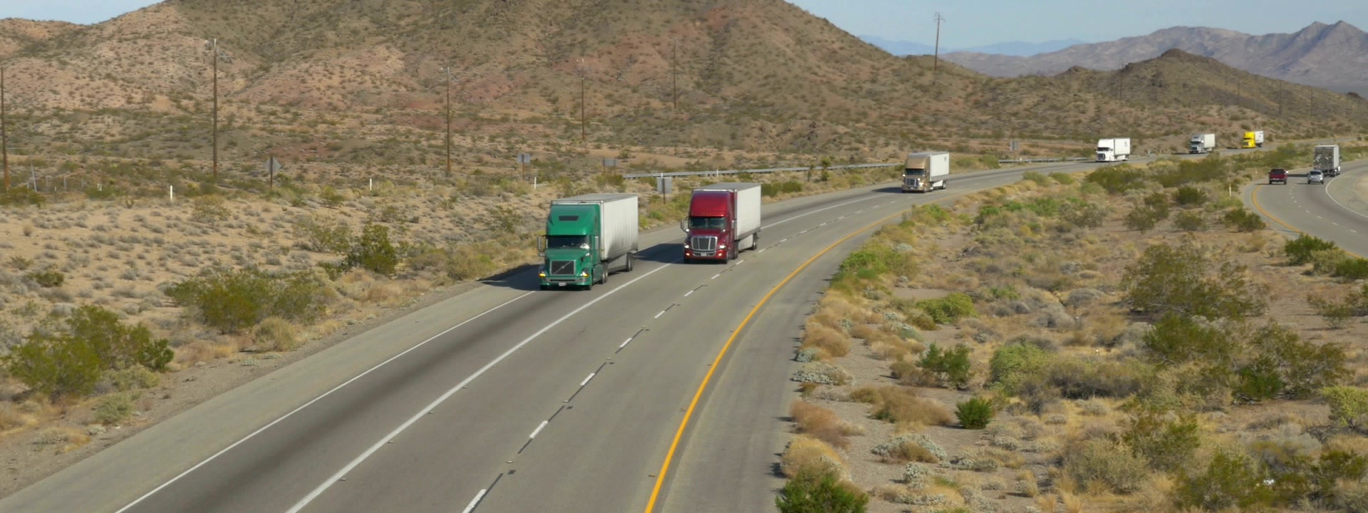 Several semi trucks driving on a highway through a desert landscape with mountains, daytime, muted earth tones